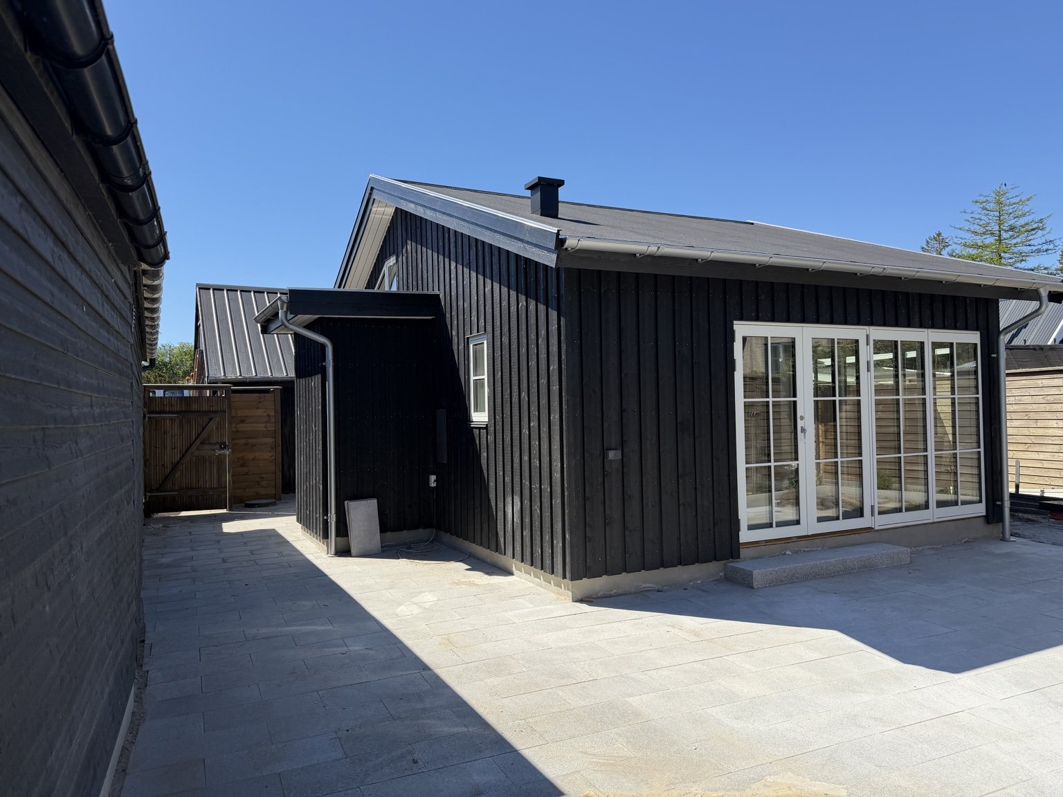 Sort sommerhus set fra hjørnet med fliseterrasse, udhus og hegn — Faxe Ladeplads
